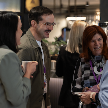 Three attendees laughing and chatting over coffee at an Amplify Alliance networking event.